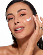 Smiling woman with bare shoulders applying a streak of white Anti-Age Lifting cream to her cheek.