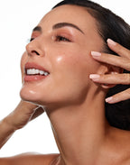 Smiling woman with radiant skin gently touching her cheek and looking upward against a white background.