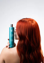 Back view of a woman with vibrant red hair holding a teal bottle of Oil Controlling Shampoo near her shoulder.