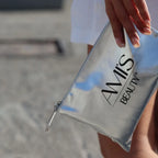 Person holding a silver metallic cosmetic bag with the Amis Beauty logo against a blurred outdoor background.
