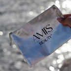 Hand holding a silver metallic cosmetic bag with the Amis Beauty logo against a backdrop of sparkling water.