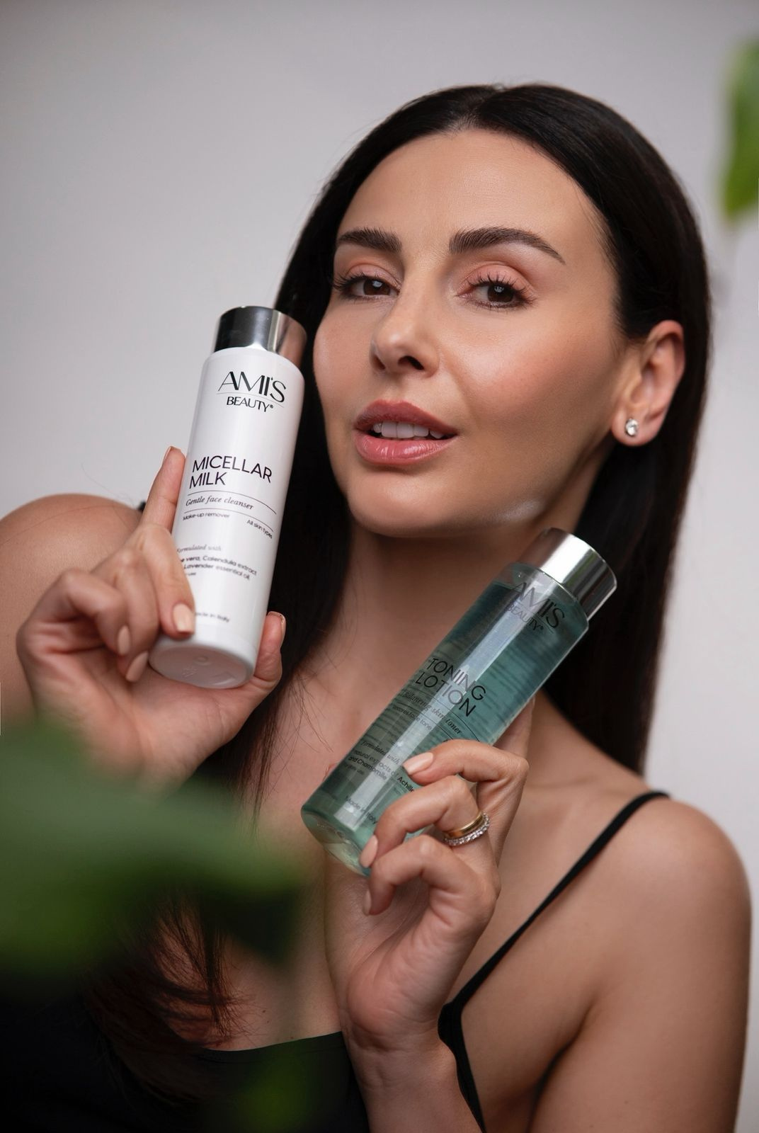 Smiling woman holding a white skincare bottle and a teal bottle of Toning Lotion, with blurred green leaves in the foreground.