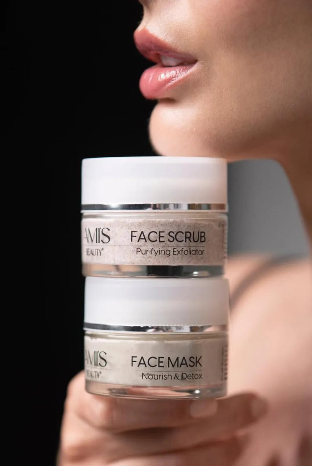 Close-up of a woman holding two stacked jars of Face Scrub Purifying Exfoliator near her face against a dark background.
