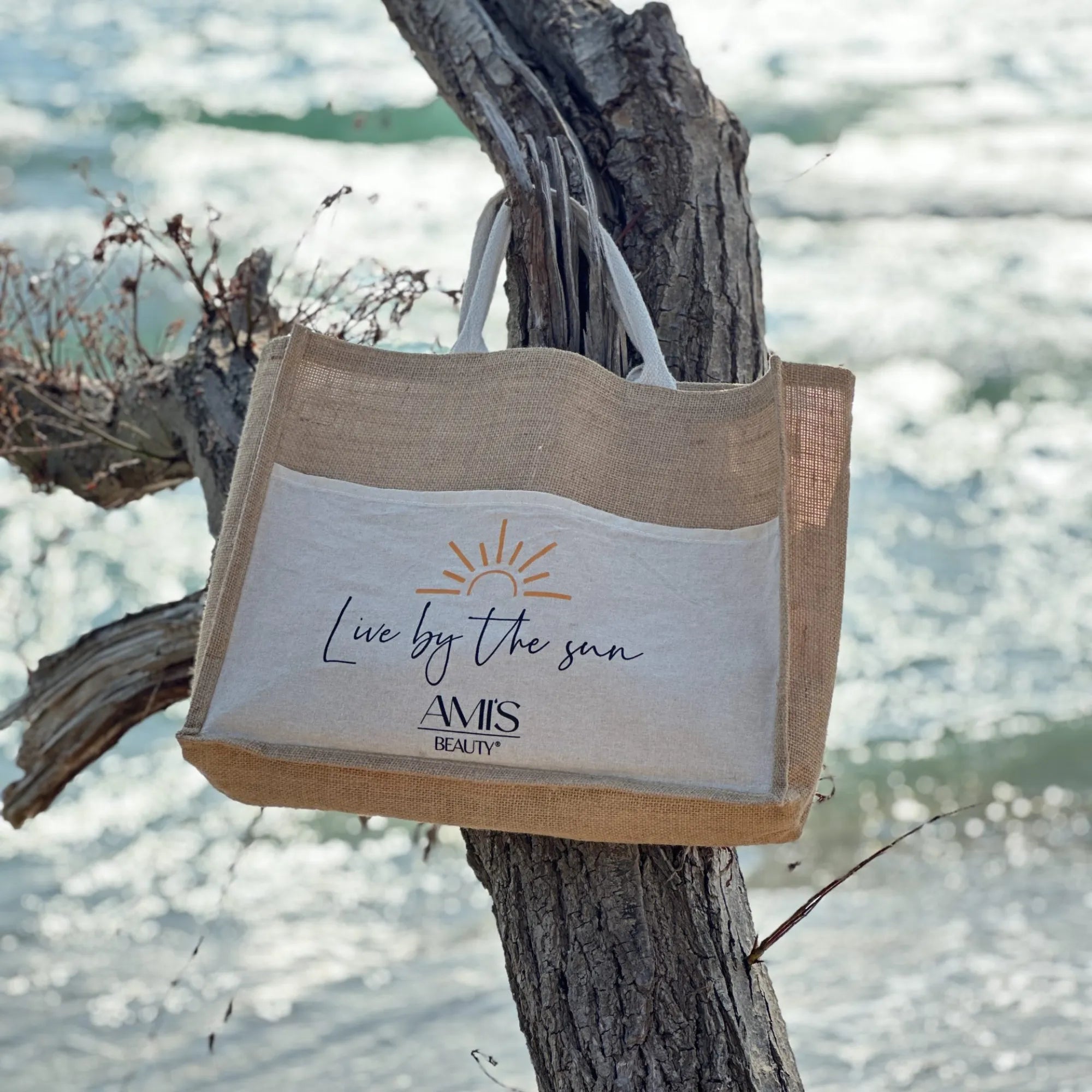 Natural woven beach tote bag with a white front panel hanging from a piece of driftwood against an ocean background.