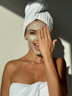 Smiling woman wrapped in towels playfully covering one eye with her hand while wearing a facial mask, standing in bright sunlight.