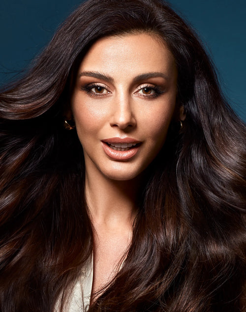 Glamour portrait of a smiling woman with voluminous, long dark wavy hair against a dark blue background.