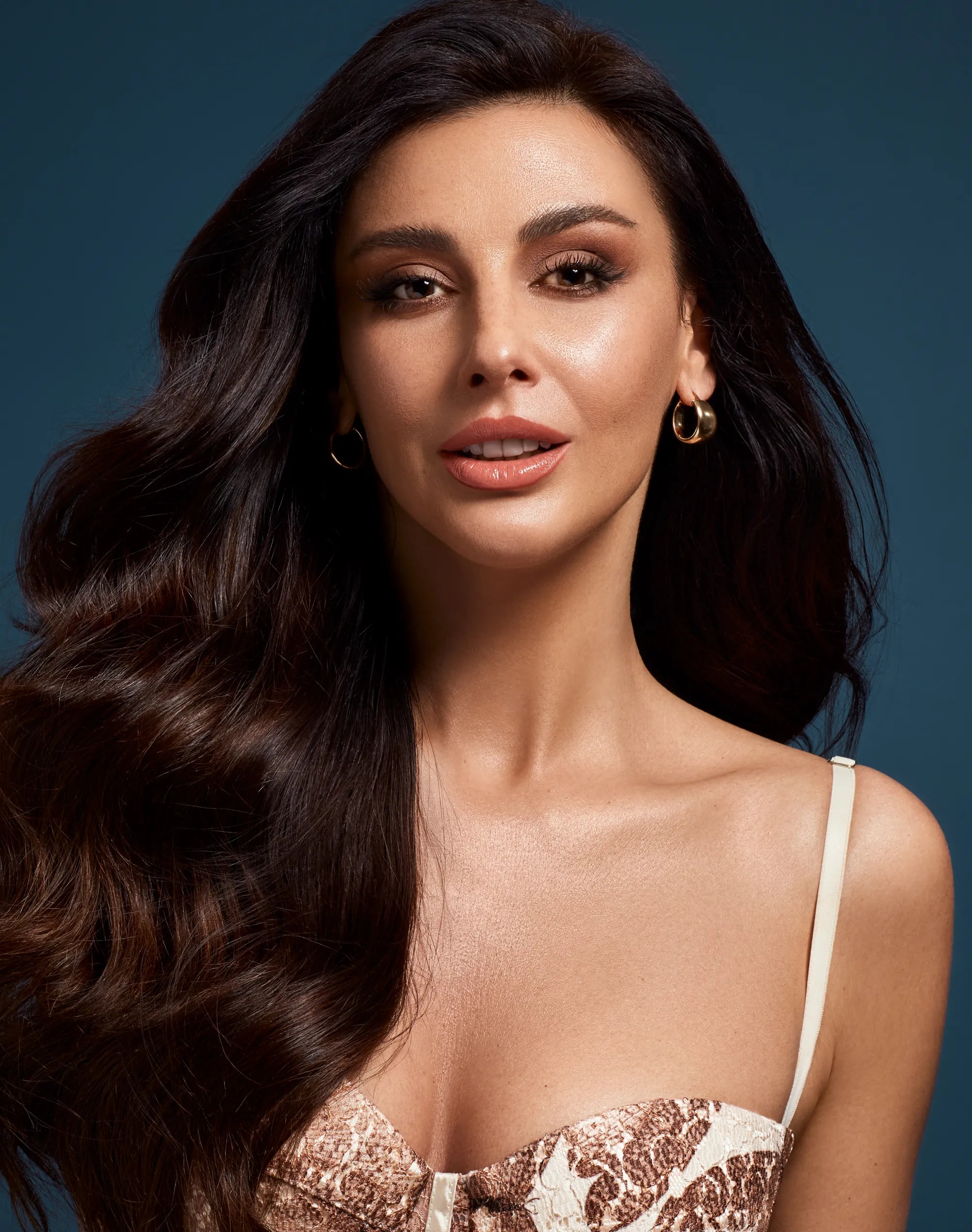 Glamour portrait of a smiling woman with voluminous, long dark wavy hair against a dark blue background.